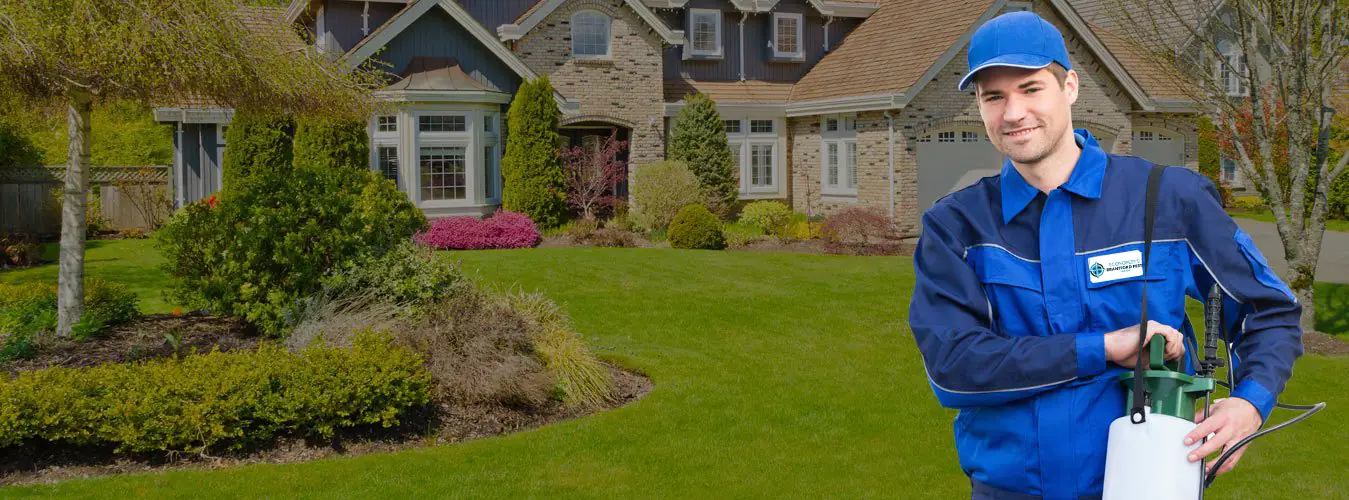 Brantford pest control technician in a blue uniform holding a pressure sprayer in front of a brick house with a green lawn